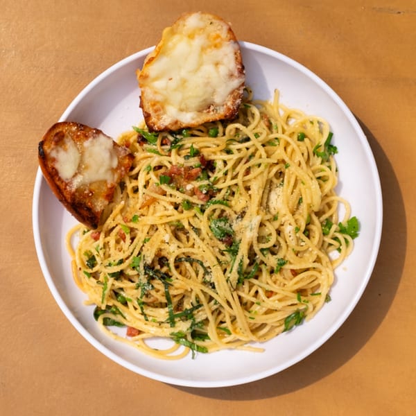 A white bowl of Tardino Brother's spaghetti with bacon, herbs, and two cheesy garlic bread slices.