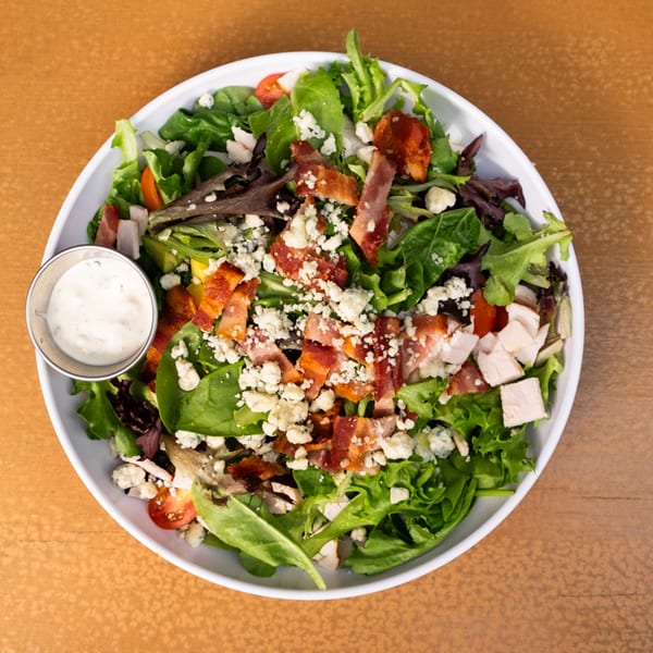 A vibrant salad with mixed greens, bacon, blue cheese, chicken, and tomatoes, served with a side of dressing.